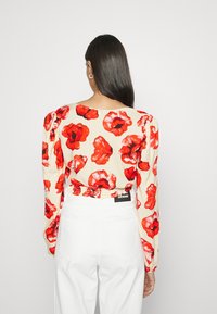 Blusa floreale a maniche lunghe con base crema e grandi motivi di papaveri rossi, vita elasticizzata e dettagli di maniche arricciate. Abbinata a pantaloni bianchi.