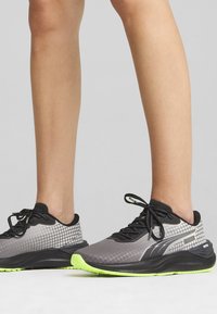 Szürke sportcipők texturált felülettel, fekete kiegészítőkkel és neon zöld talppal. Rácsos mintázattal és fekete fűzőkkel rendelkezik.
