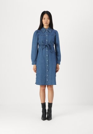 Robe chemise en denim bleu avec manches longues, patte de boutonnage à l'avant, taille ceinturée, fentes latérales et détails de coutures contrastées. Associée à des bottines noires.
