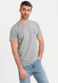 Camiseta gris de manga corta con pequeñas motas negras, cuello redondo y un gráfico circular en el área del pecho izquierdo. Textura suave.