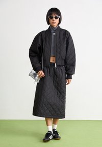 Svart quiltad bomberjacka med ribbade muddar, grå crop top, lång svart quiltad kjol och svarta sportskor med vita detaljer. Silverfärgad handväska.