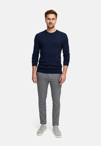 Granatowy sweter dżersejowy z teksturowanym wzorem, szare spodnie o dopasowanym kroju i jasnoszare sneakersy. Model stoi na jednolitym białym tle.