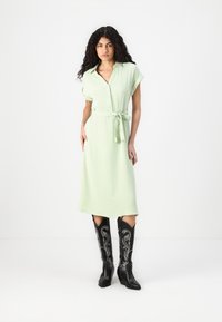 Rochie verde deschis cu mâneci scurte, închidere cu nasturi în față, talie legată. Asortată cu cizme cowboy negre cu broderie florală albă.