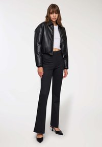 Kobieta ubrana w czarną skórzaną kurtkę typu cropped, białą bluzkę cropped, czarne spodnie w paski oraz czarne szpilki, stojąca na tle o jednolitej barwie.