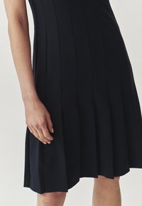Navyblaues plissiertes Kleid mit tailliertem Oberteil, schwingendem Rock und glatter Stoffstruktur. Nahaufnahme einer Hand, die auf dem Rock ruht.