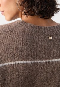 Brauner Strickpullover mit strukturierter Oberfläche, ausgestattet mit einem weißen horizontalen Streifen und einem kleinen goldenen LOGO-Tag auf der Rückseite.