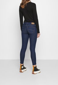 Jeans ajustados de color azul con una cintura alta y una etiqueta de logo beige en la parte posterior. Combinados con zapatos en blanco y negro con detalles en naranja.