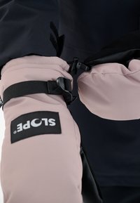 Les manchons isolés roses présentent une sangle noire et une boucle pour l'ajustement, avec une étiquette de logo noire bien visible sur le tissu. L'arrière-plan est sombre.