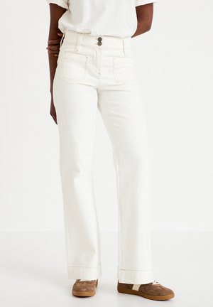 Osoba nosząca białe jeansy z wysokim stanem, z przednimi naszywanymi kieszeniami oraz brązowe trampki, stojąca na tle jednolitego białego tła.