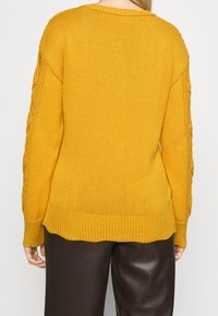 Senapsgul tröja med ribbad nederdel och muddar. Har ett stickat mönster längs ärmarna. Texturerad tyg ger ett mysigt utseende.