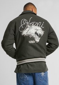 Dark green jacket with a large white tiger embroidery on the back, featuring barbed wire accents and a striped hem.
