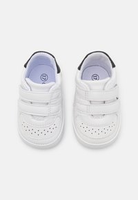 Białe buciki dla niemowląt z dwiema rzepami, zaokrąglonym noskiem i perforowanymi detalami. Czarne akcenty na pięcie oraz miękka podszewka wewnętrzna.