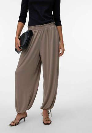 Kvinde iført løse taupe bukser med stramme ankler, sort bluse med trekvartærmer, holder sort clutch og har hælede sandaler på.