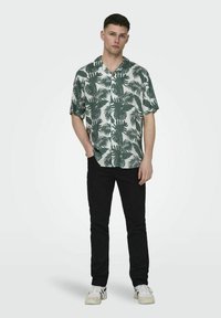 Cămașă cu mânecă scurtă, verde și albă, cu un model de frunze de palmier, combinată cu pantaloni negri și adidași albi. Textura țesăturii este netedă.