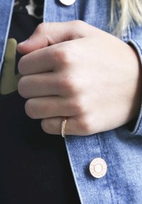 Roze gouden ring met kleine heldere stenen, gedragen op een licht gebruinde hand. Een spijkerjack met een metalen knop wordt op de achtergrond getoond.