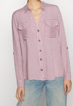 Kobieta nosząca różowo-białą pionowo prążkowaną koszulę na guziki z długim rękawem, z dwoma przednimi kieszeniami na klapę i jasnoniebieskie jeansy.