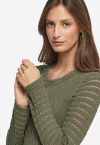 Grön stickad tröja med långa ärmar som har öppna stickmönster, rund halsringning och strukturerad textil. Inga ytterligare detaljer.