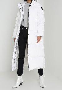 Lungo cappotto imbottito bianco con cappuccio, caratterizzato da un accento nero sulla spalla, abbinato a jeans neri e stivaletti bianchi con tacco.
