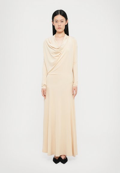 Robe en jersey longue beige avec un décolleté drapé et des manches longues. Texture lisse, ajustée au corsage, et évasée vers l'ourlet. Chaussures noires.