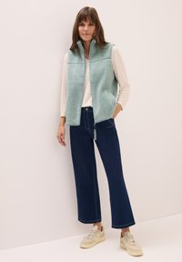 Ljus turkos fleeceväst med hög krage, framdragkedja, två sidofickor, i kombination med marinblå vida jeans och beige sneakers.