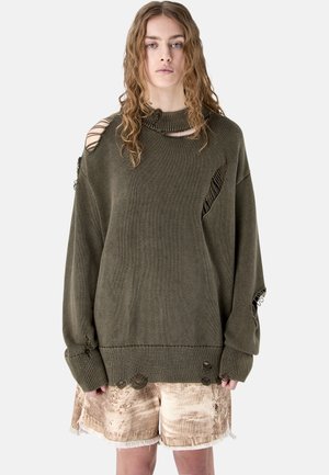 Donna con lunghi capelli ricci che indossa un ampio maglione verde scuro usurato e pantaloncini beige con bordi sfrangiati, in piedi davanti a uno sfondo bianco.