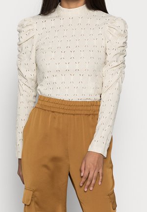 Femme portant un chemisier texturé crème à manches longues avec épaules bouffantes et un pantalon moutarde taille haute avec poches latérales.