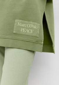 Felpa verde con uno spacco laterale, dotata di un'etichetta rettangolare con la scritta "Marc O'Polo PEACE" in carattere chiaro. Tessuto morbido e testurizzato.