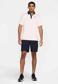 Jasno beżowa koszulka polo z granatowym kołnierzykiem, zestawiona z granatowymi szortami. Model ma na sobie czarne buty sportowe. Prosty design z krótkimi rękawami.