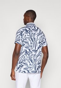 Camisa polo de manga curta em branco com padrão abstrato de ondas azul marinho. Apresenta um colarinho clássico e um design slim fit. Tecido suave e leve.