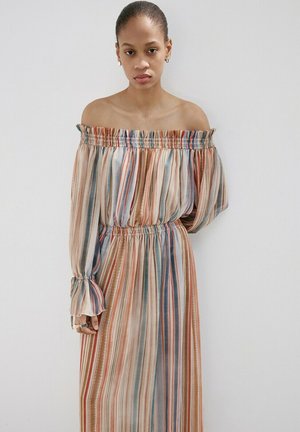 Femme portant une longue robe épaules dénudées avec des rayures verticales multicolores dans des tons de bleu, orange et beige sur un fond uni.