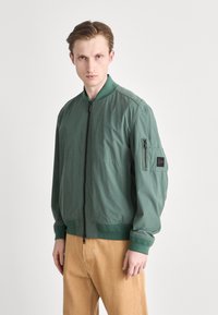 Bunda ve stylu bomber v tmavě zelené látce s předním zipem, žebrovanými manžetami a náprsní kapsou s logem značky. Hladký povrch.