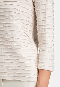 Hellbeige Oberteil mit einem strukturierten Wellenmuster, ausgestattet mit Dreiviertelärmeln und einem weichen, dehnbaren Stoff.