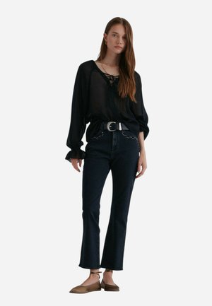 Zwarte sheer blouse met lange mouwen en vetersluiting, gecombineerd met donkerdenim flared jeans met studversieringen en een zilveren riem.