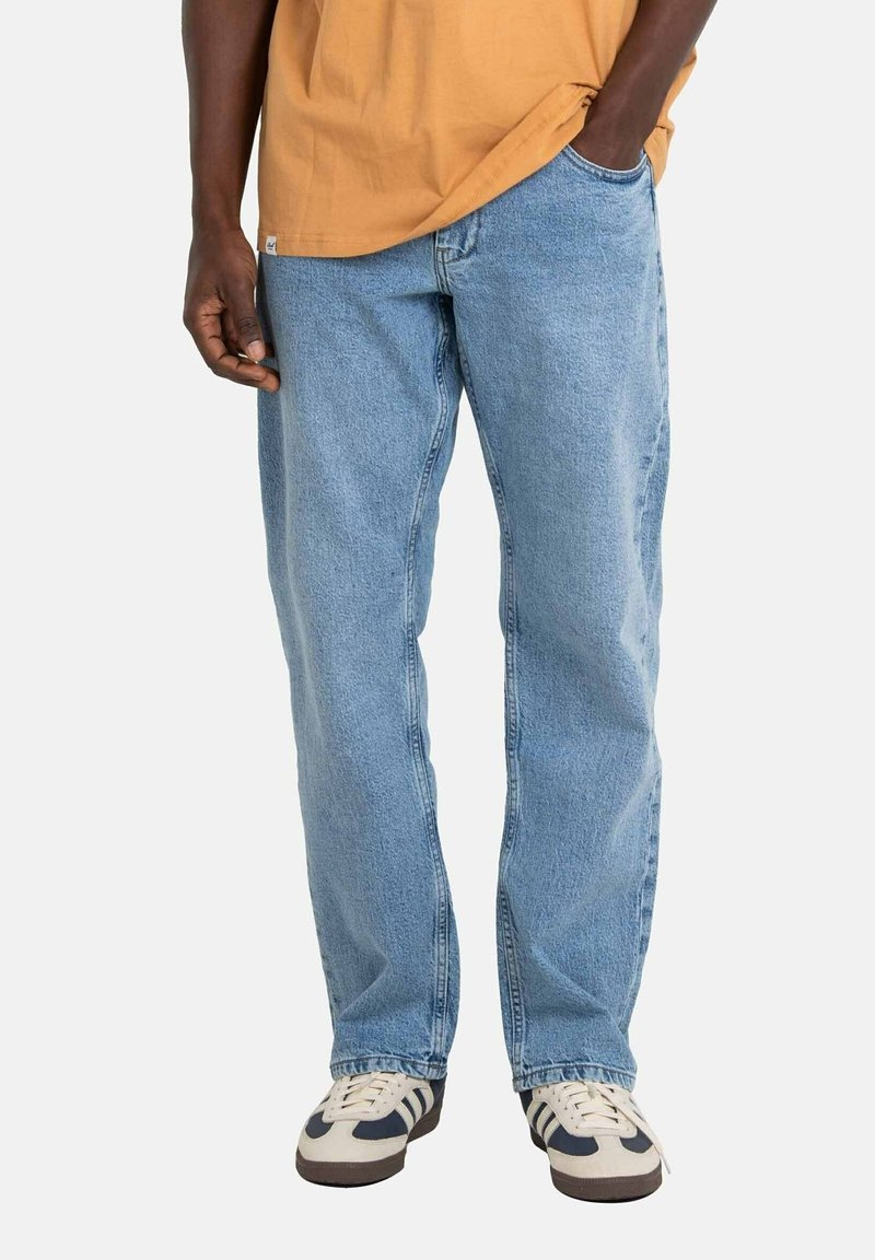 Homme portant un jean en denim bleu clair, des baskets beige et noir, et un t-shirt orange avec une main dans la poche.