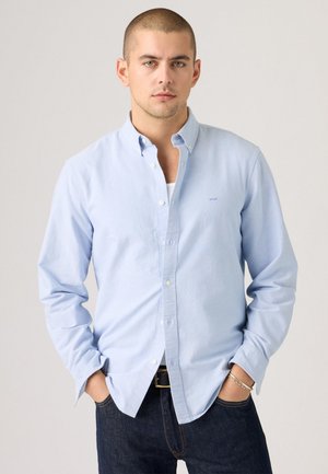 Camisa de botón azul claro con mangas largas, cuello estándar y un sutil logotipo de la marca. Se lleva con jeans oscuros y un cinturón negro.