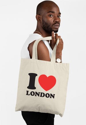Uomo che indossa una maglietta senza maniche bianca tiene una borsa beige con il testo "I ❤️ LONDON" in nero e rosso su sfondo bianco semplice.