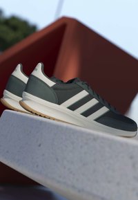 Chaussures de sport gris foncé avec des rayures blanches et des semelles intermédiaires blanches reposant sur une surface en béton clair contre un fond géométrique brun rouille flou.