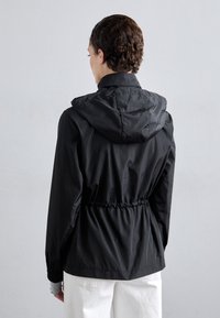 Person som bär en svart huva jacka med insvängd midja och elastiska ärmslut, kombinerad med vita byxor, vänd bort från kameran mot en enfärgad bakgrund.