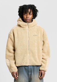 Beige fleecejacka med huva, med dragkedja i fronten, texturerad tyg och kontrasterande släta tygdetaljer på ärmarna.