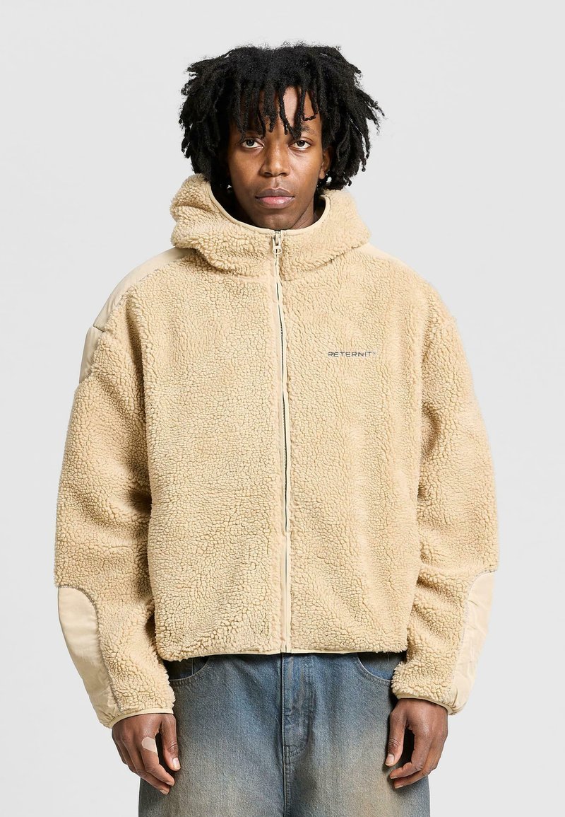 Beige fleecejacka med huva, med dragkedja i fronten, texturerad tyg och kontrasterande släta tygdetaljer på ärmarna.