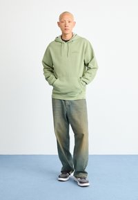 Πράσινο φούτερ με μπροστινή τσέπη και κορδόνια, συνδυασμένο με χαλαρά μπλε τζιν. Το μοντέλο φοράει γκρίζα και λευκά αθλητικά παπούτσια σε μπλε δάπεδο.