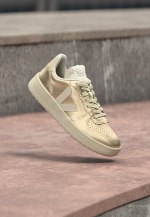 Baskets décontractées beige et dorées avec lacets et logo sur le côté, suspendues au-dessus d'une surface en pierre rose clair avec un fond en pierre grise.