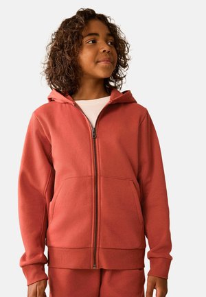 Child with curly hair wearing a rust-colored zip-up hoodie and matching pants looking to the side on a plain background.