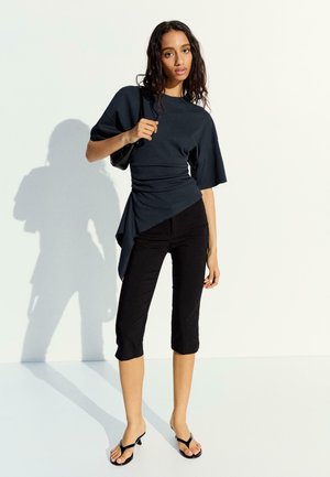 Junge Frau, die ein dunkles asymmetrisches Oberteil, kurze schwarze Hosen, schwarze Sandaletten mit Absatz trägt und eine kleine schwarze Umhängetasche hält, steht in Innenräumen.