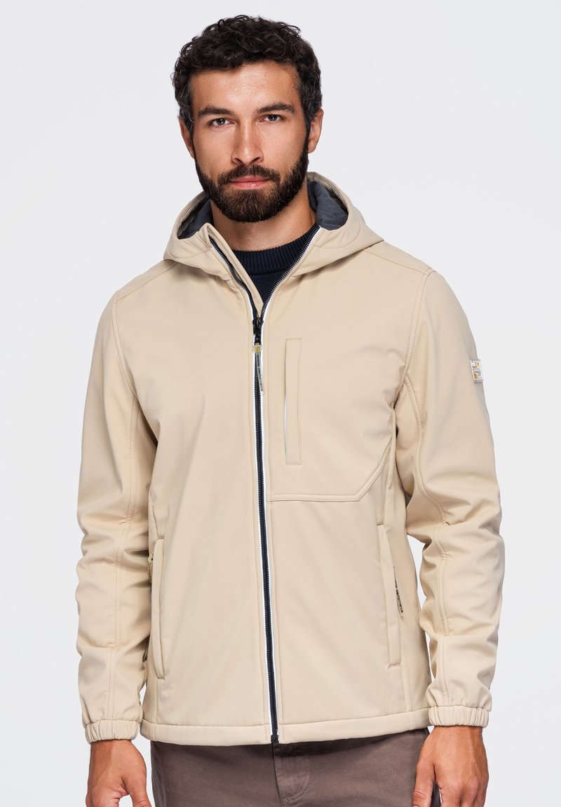 Homme aux cheveux bouclés et foncés avec une barbe, portant une veste à capuche beige zippée sur une chemise sombre, debout sur un fond clair uni.