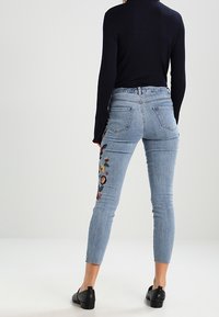 Jeans ajustados de color azul claro con bordado floral en el lado izquierdo, cinco bolsillos y una cintura de altura media, combinados con una blusa de punto azul marino.