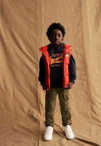 Gilet rembourré rouge porté sur un sweat à capuche noir avec impression orange, associé à un pantalon cargo olive et des baskets blanches. Textures lisses et couches variées.