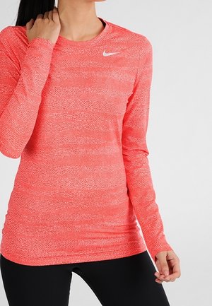 Kobieta w dopasowanej, fakturowanej różowej koszulce z długim rękawem Nike oraz czarnych spodniach sportowych, stoi na tle jasnego, jednolitego tła.