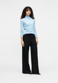 Ανοιχτό μπλε μπλούζα με μακριά μανίκια και ζιβάγκο, με μικρό λογότυπο στο στήθος, σε συνδυασμό με μαύρο παντελόνι φαρδύ και μυτερά μαύρα παπούτσια.