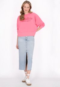 Felpa oversized rosa con la scritta "Happiness Invisibile", abbinata a una gonna di jeans azzurra chiara con uno spacco frontale. Sneakers bianche completano l'outfit.